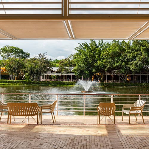 Cuatro sillas y un sillón alargado color beige en frente de un lago, en el que hay una fuente con muchos árboles alrededor y una instalación perteneciente al hotel Lagosol